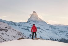 אדם במעיל אדום עומד על הר מושלג, בוהה אל עבר האופק המושלג בזמן שקיעה חושב על איך להגיע לפסגה